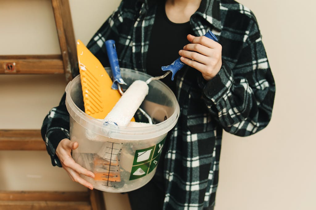 A person holds a bucket containing a paint roller and scraper for a renovation project.